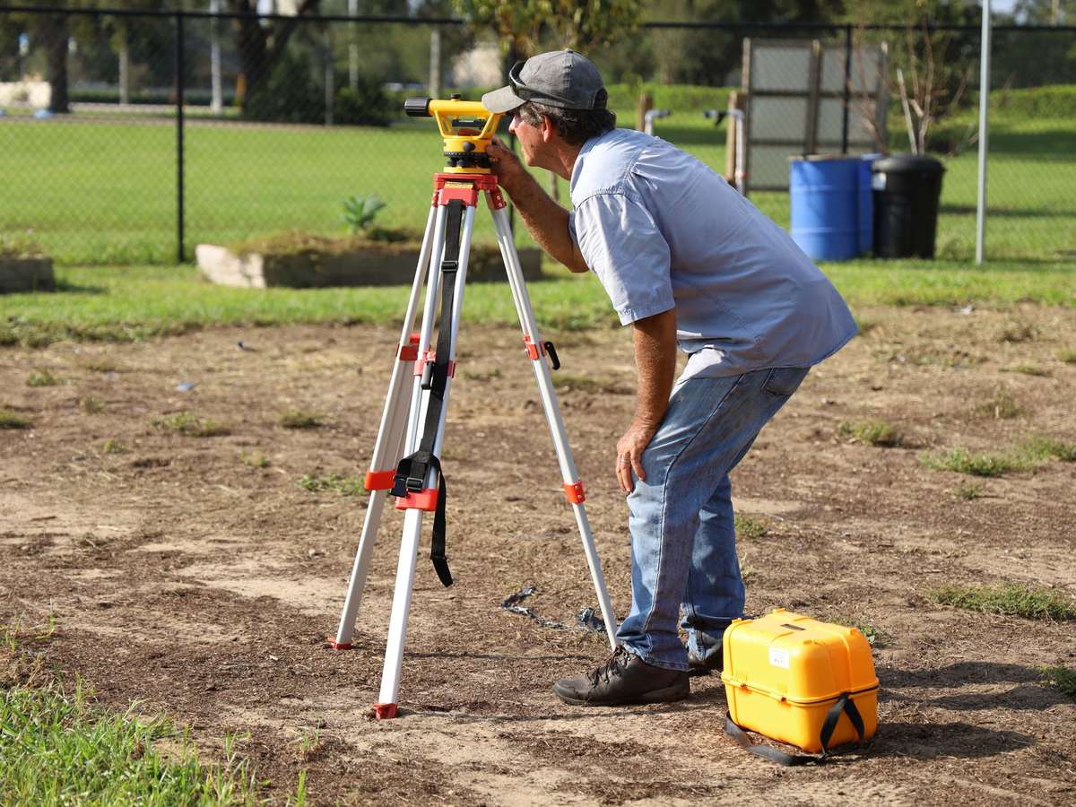 A man in a gray cap and jeans leans to look through a yellow automatic level on a tripod on bare dirt, with a chain-link fence behind him