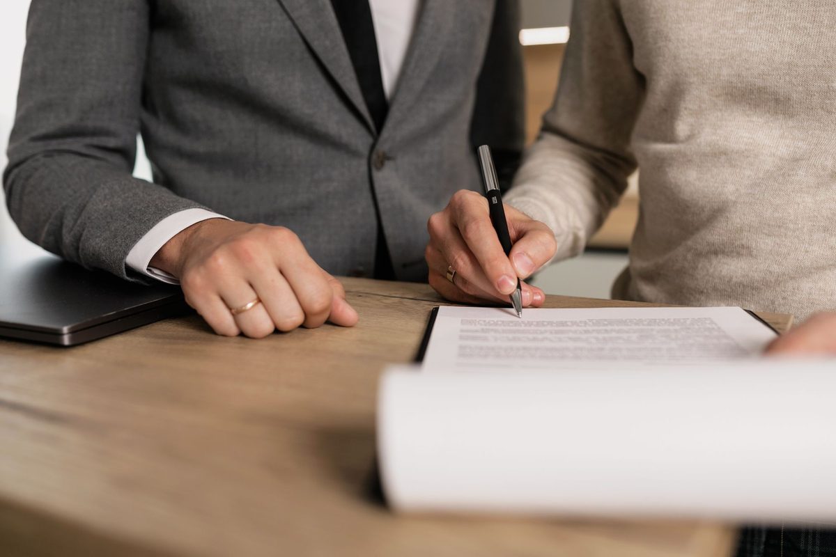 Close-up of two people at a wooden table: one in a gray suit jacket rests a hand beside a closed laptop while the other, in a beige sweater, signs a printed document on a clipboard
