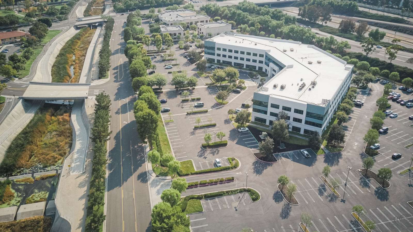 Aerial view of a multistory office building surrounded by striped surface parking lots beside a divided road