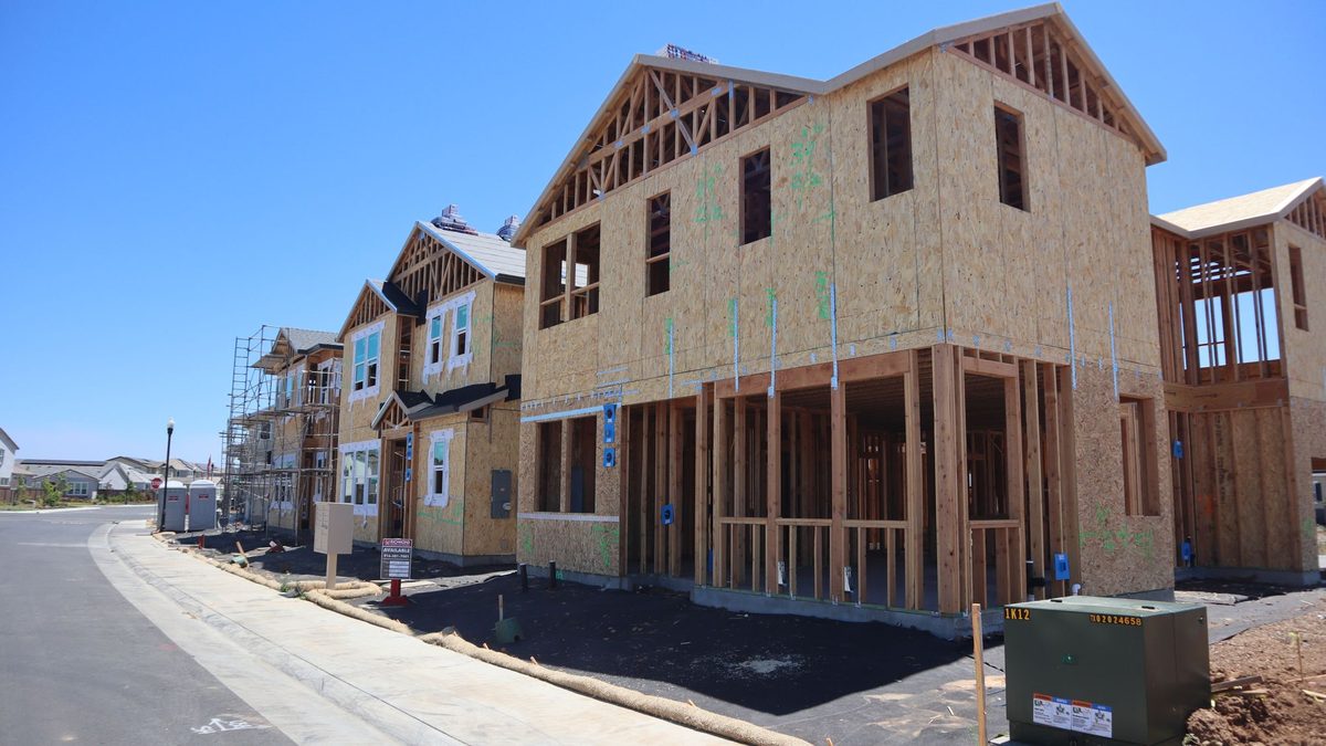 A row of wood-framed houses under construction along a new curving street, with tan fiber rolls laid along the back of the sidewalk and a green utility box in the foreground, under a clear blue sky