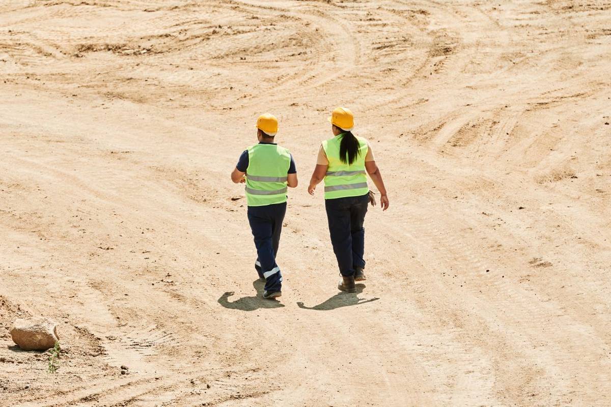 Seen from behind, two workers in yellow hard hats and high-visibility vests walk across a large graded dirt site marked with tire tracks