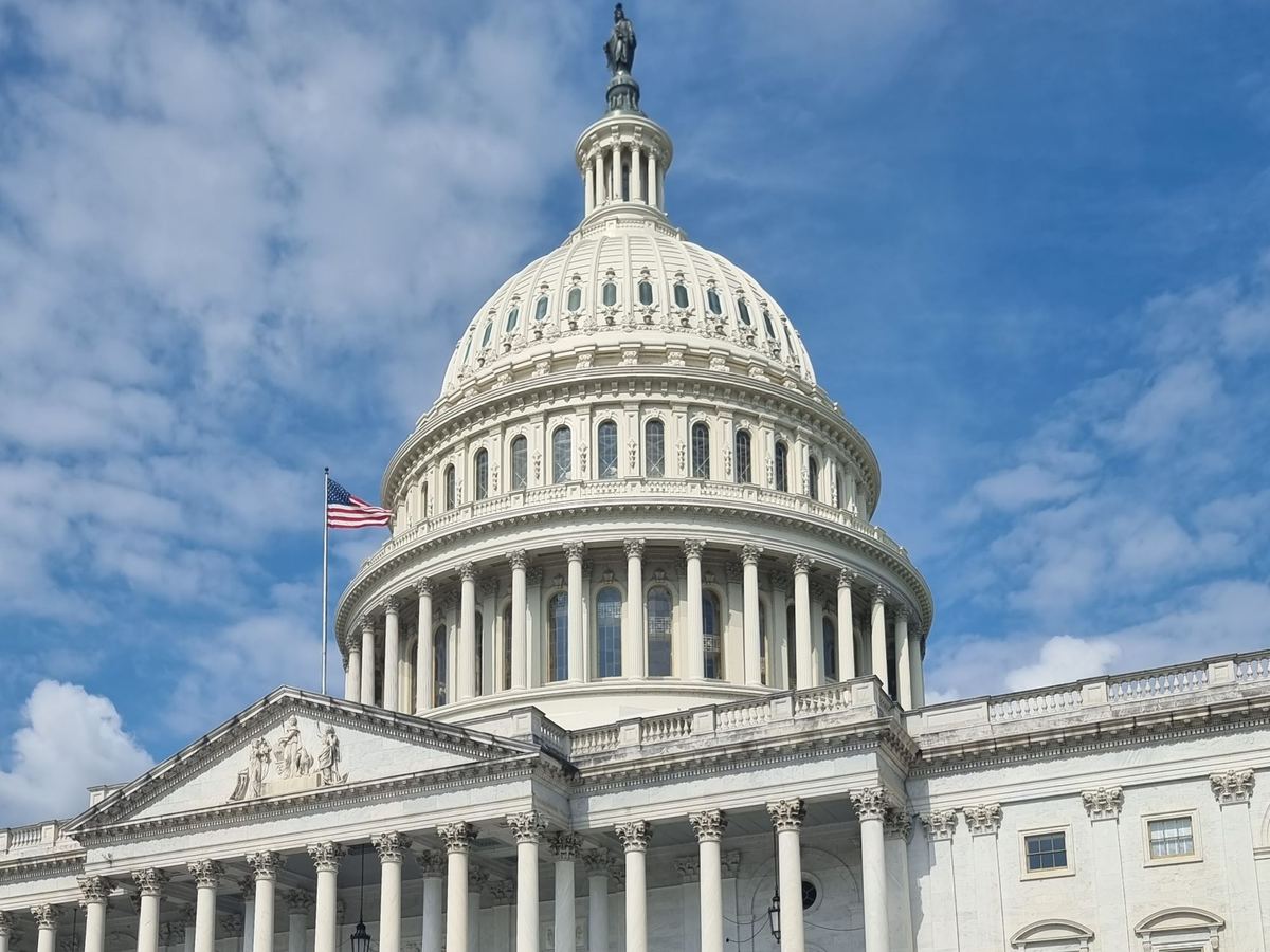 The dome of the U.S. Capitol against a blue sky with white clouds, with an American flag flying at left above the columned facade
