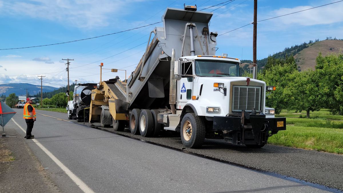 A white dump truck with its bed raised feeds a paving machine on a two-lane rural road, while a worker in an orange high-visibility vest stands at the roadside