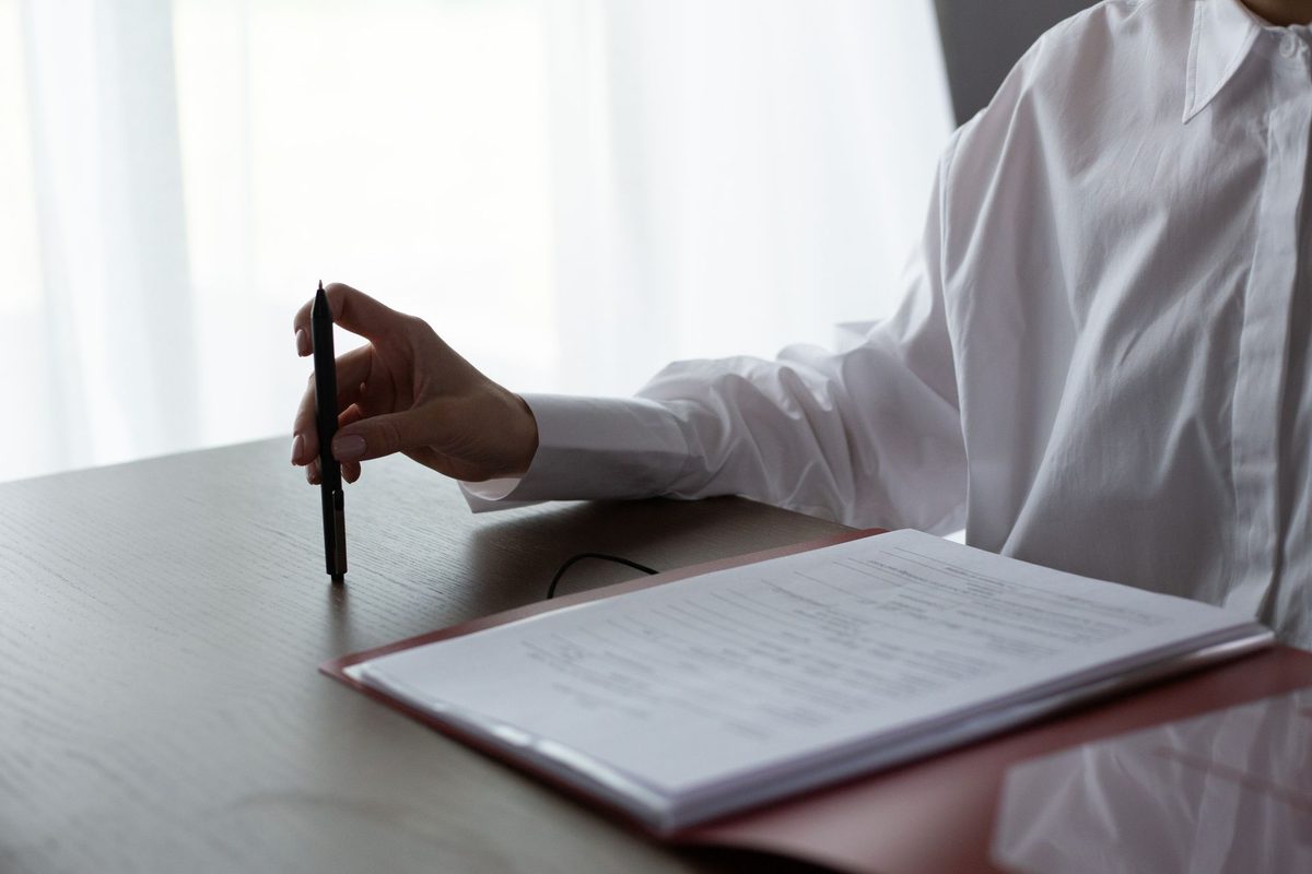 A person in a white shirt, face out of frame, holds a pen upright above a dark wooden desk beside an open red folder of printed documents