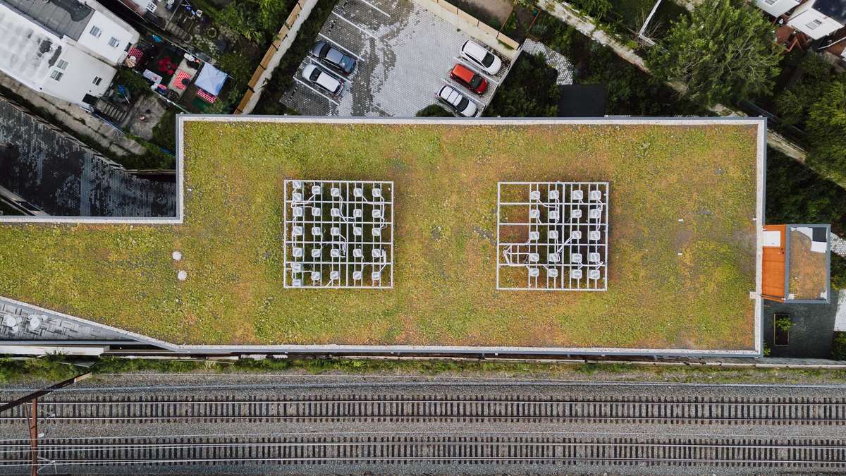 Aerial view looking straight down on a flat building roof covered in low green and tan plants, with two white frames holding rooftop air-conditioning units, a small parking area above and railroad tracks below