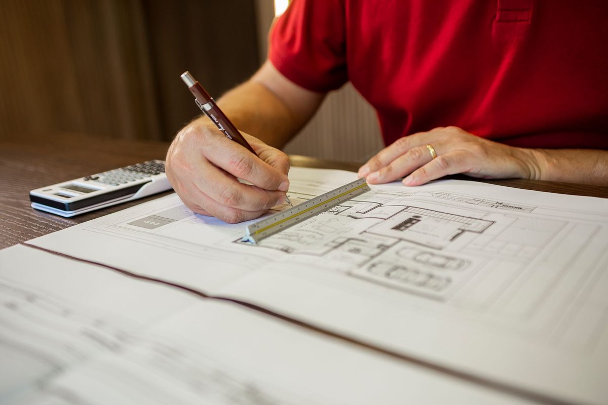 A man in a red polo shirt draws on a printed architectural plan with a mechanical pencil beside a triangular scale ruler, with a calculator on the wooden desk