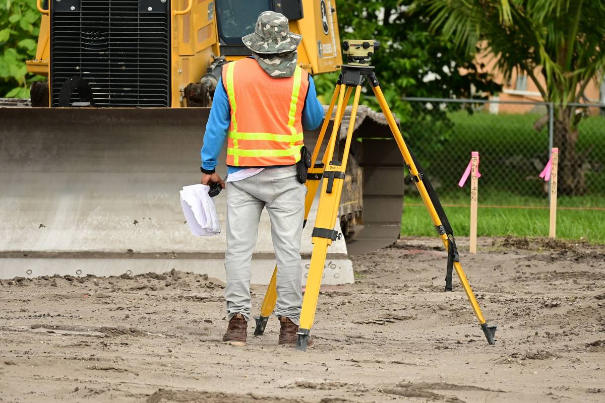 A surveyor in an orange high-visibility vest and a camouflage hat, seen from behind, stands beside an automatic level on a yellow tripod on a graded dirt lot, holding papers, with wooden stakes with pink flagging and a yellow bulldozer nearby