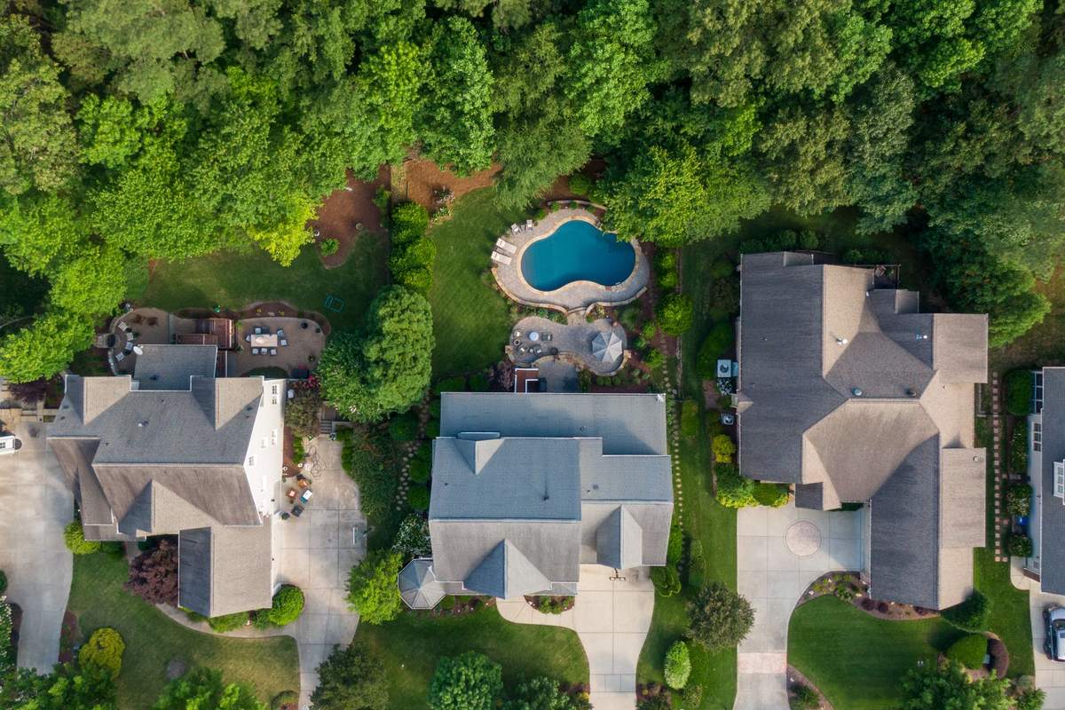 Top-down aerial view of neighboring houses with gray roofs, driveways, lawns, backyard patios and a swimming pool, backed by dense green trees