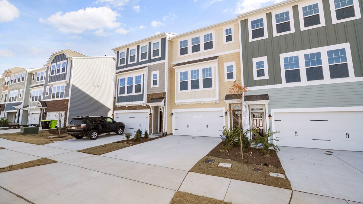 A row of new three-story townhouses with white garage doors, short concrete driveways and young landscaping along a sidewalk under a partly cloudy sky