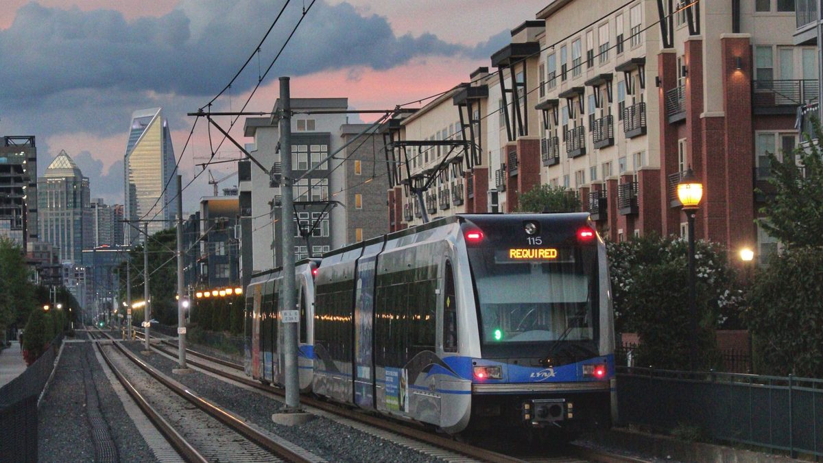 A two-car silver-and-blue light rail train runs on tracks beside multistory apartment buildings with balconies, under overhead wires, with a city skyline against a pink dusk sky in the distance