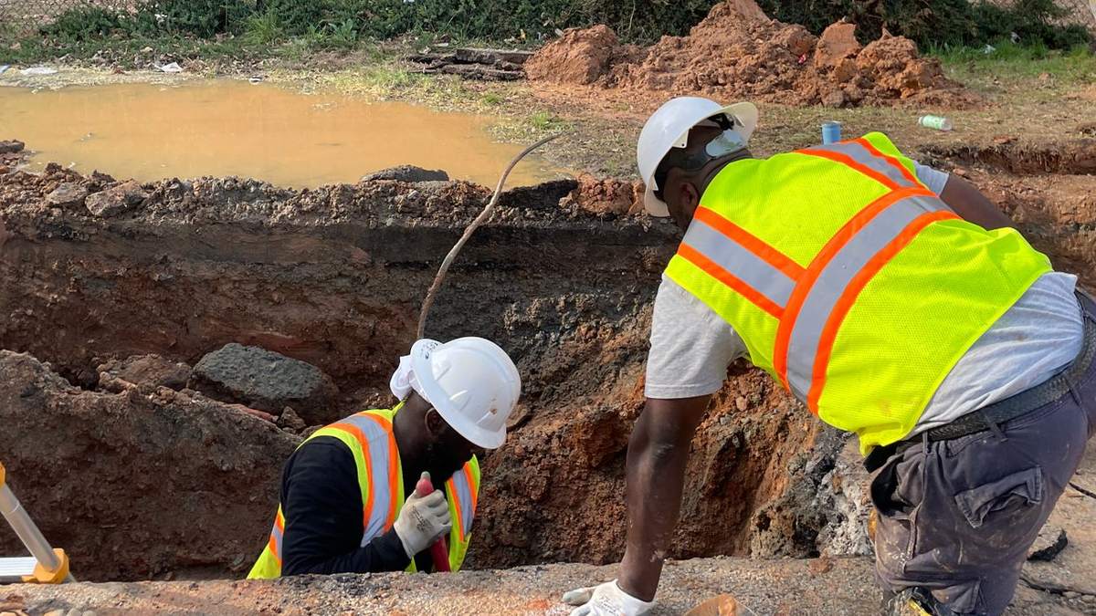 Two construction workers in white hard hats and high-visibility vests, one standing in an excavated utility trench and one leaning over its edge, next to a muddy pool of water and a pile of red soil