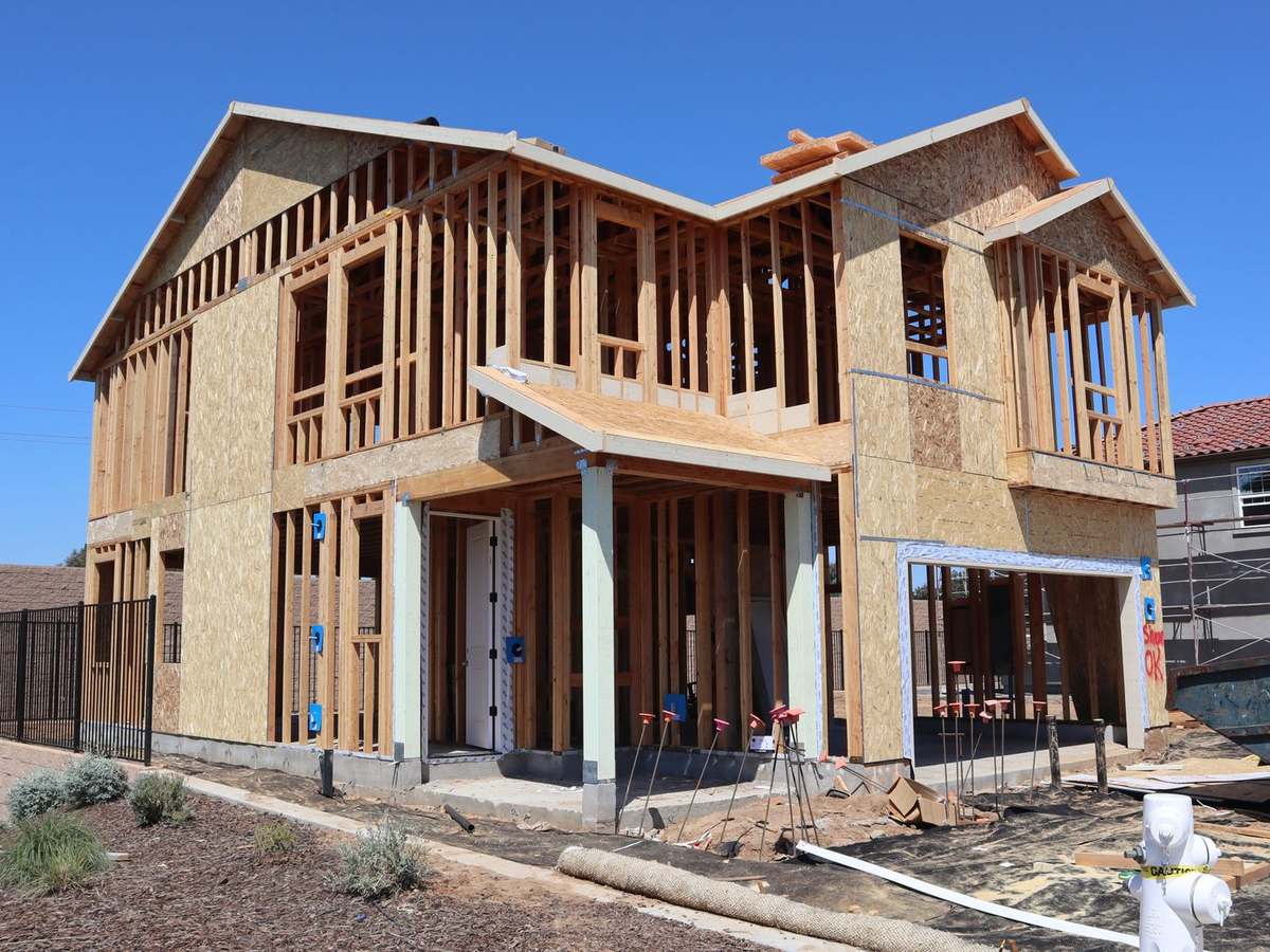 A two-story wood-framed house under construction, with sheathing on part of the walls and open framing on the upper floor, a black metal fence at left and a white fire hydrant in front, under a clear blue sky