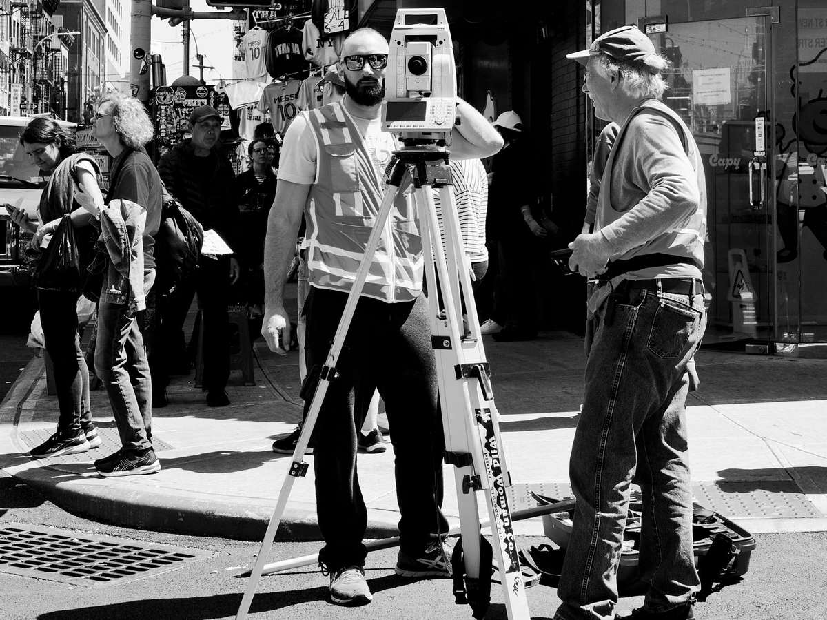Black-and-white photo of a surveyor in a high-visibility safety vest and sunglasses standing behind a total station on a tripod at a city street corner, with an older man in a cap beside him and pedestrians and shops behind them