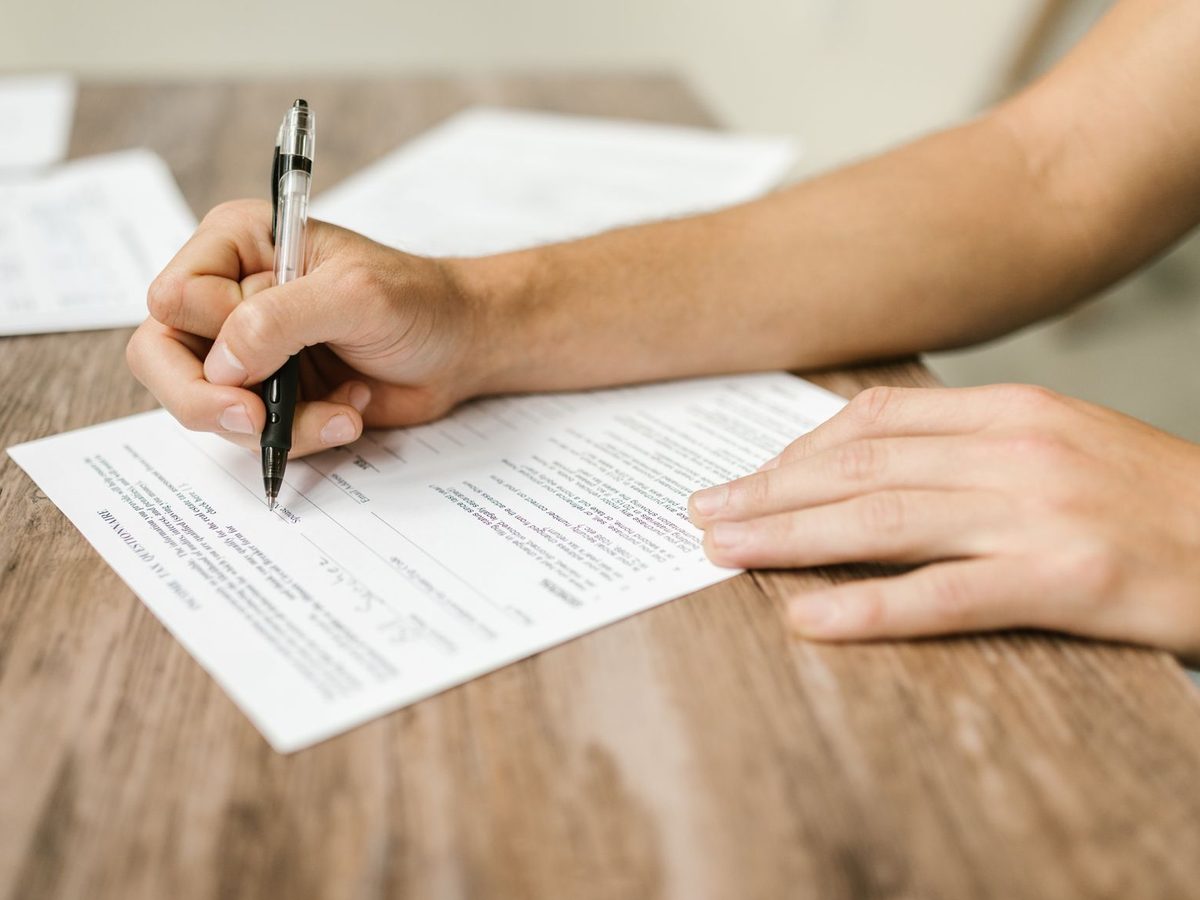Close-up of a hand holding a black pen and filling in a printed paper form on a wooden desk, with other papers in the background