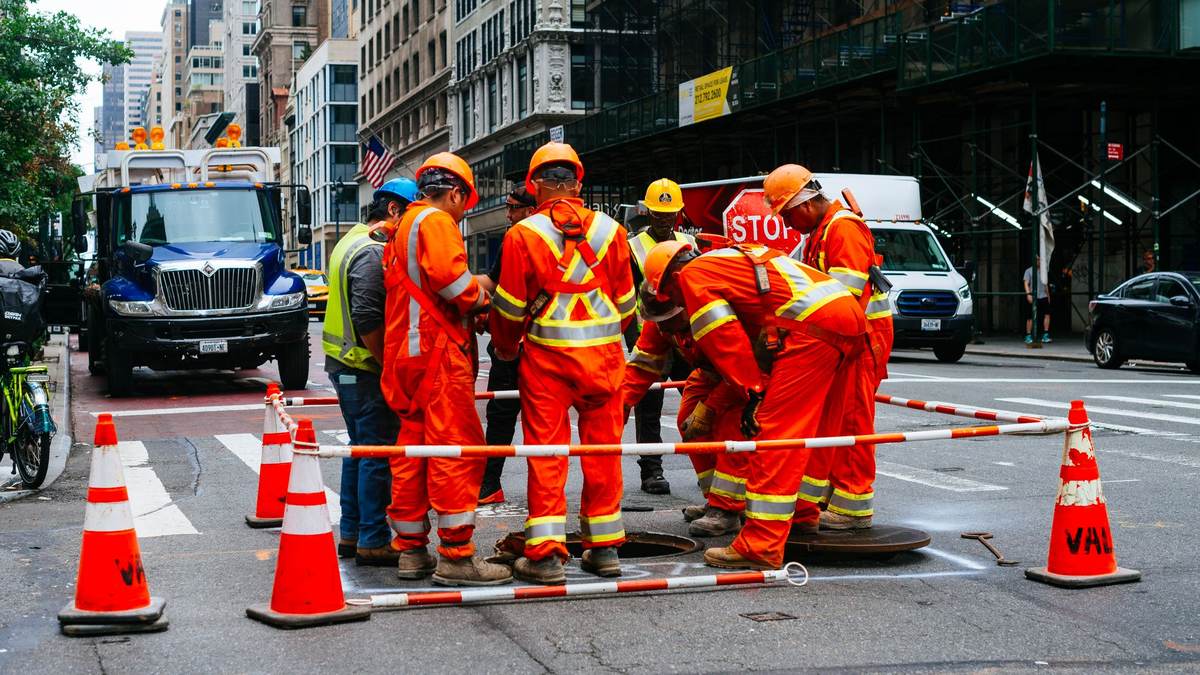 A street crew in hard hats and orange and yellow high-visibility workwear gathers around an open manhole inside a ring of traffic cones joined by red-and-white striped poles, with a blue truck and tall city buildings behind