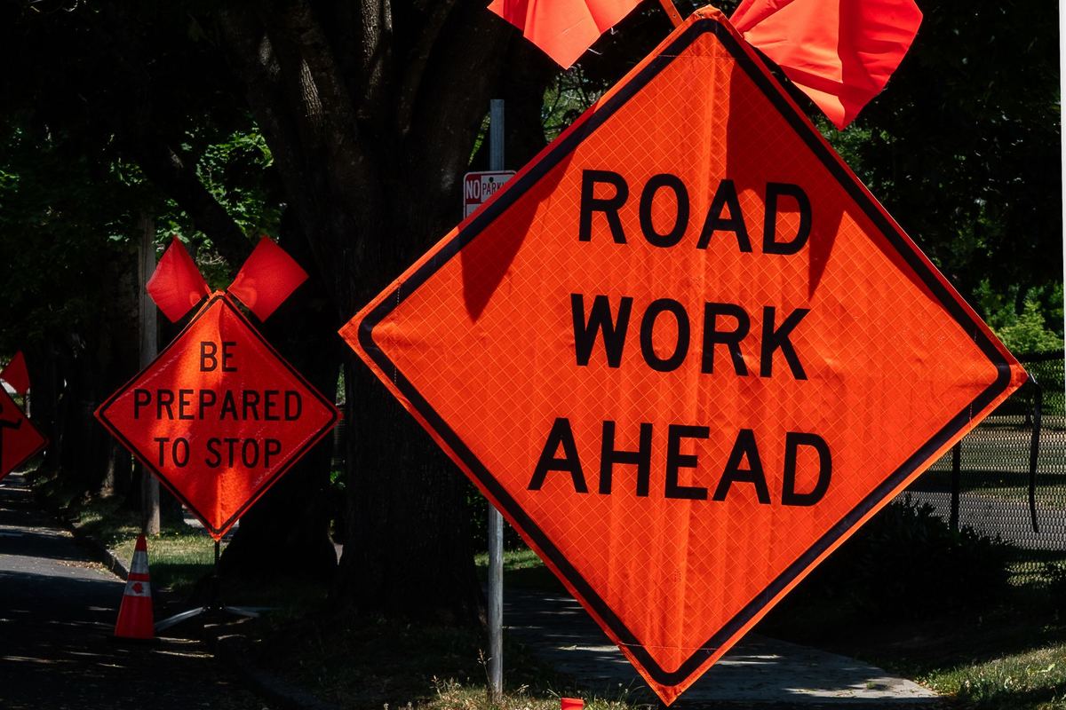 Orange diamond-shaped ROAD WORK AHEAD and BE PREPARED TO STOP signs topped with orange flags, with a traffic cone, on a shaded, tree-lined street