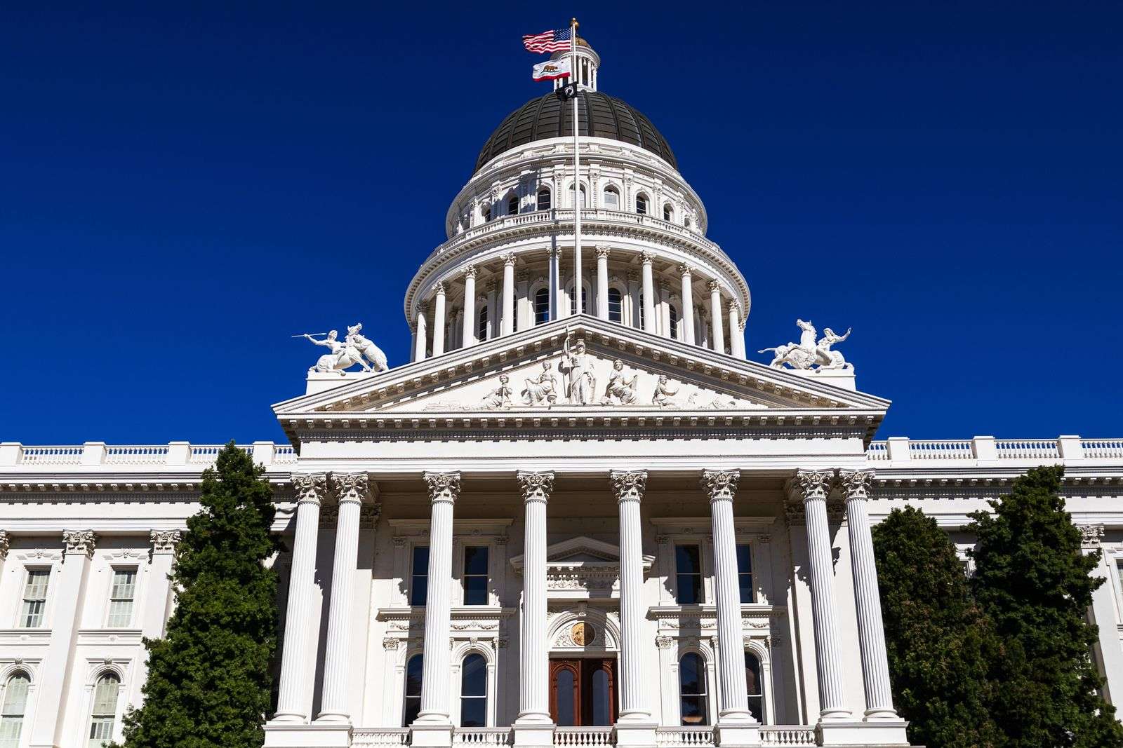 The California State Capitol in Sacramento under a clear blue sky, with the U.S., California, and POW/MIA flags flying above the dome