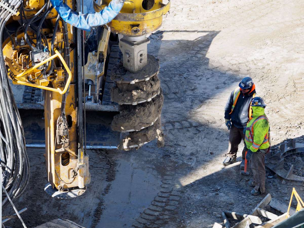 A yellow drilling rig lifts a large steel auger caked with soil out of the ground while two workers in hard hats and high-visibility vests stand nearby on a dirt site