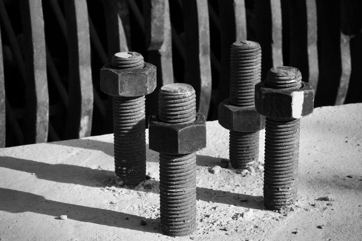 Black-and-white close-up of four threaded steel bolts with hex nuts projecting from a concrete surface in front of a dark metal grille