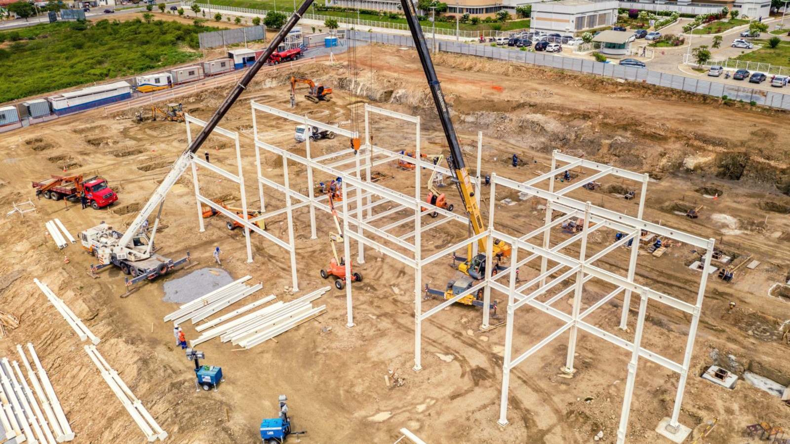 Aerial view of a steel building frame being erected with mobile cranes on a graded dirt construction site