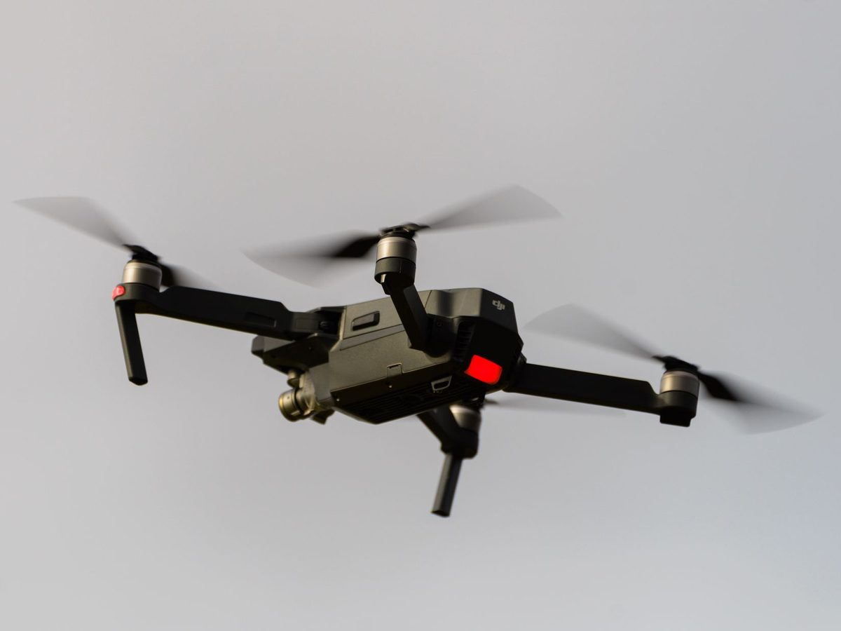A black quadcopter drone with a camera mounted under its body flies against a pale gray sky, its four propellers blurred by motion