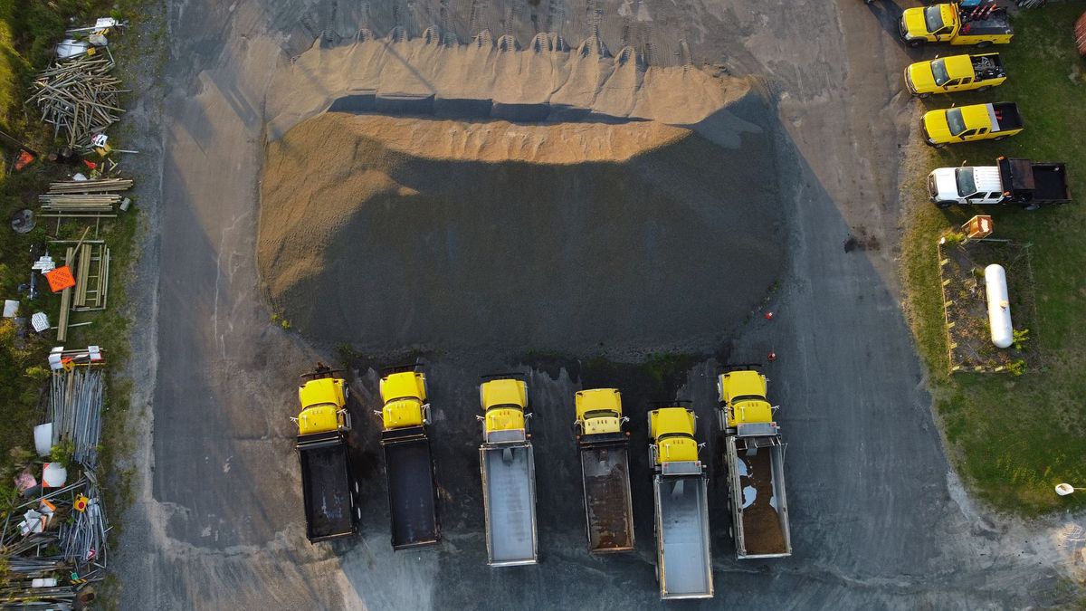 Straight-down aerial view of a large gravel stockpile in a paved yard, with six yellow dump trucks parked in a row below it and yellow pickup trucks on the grass to the right