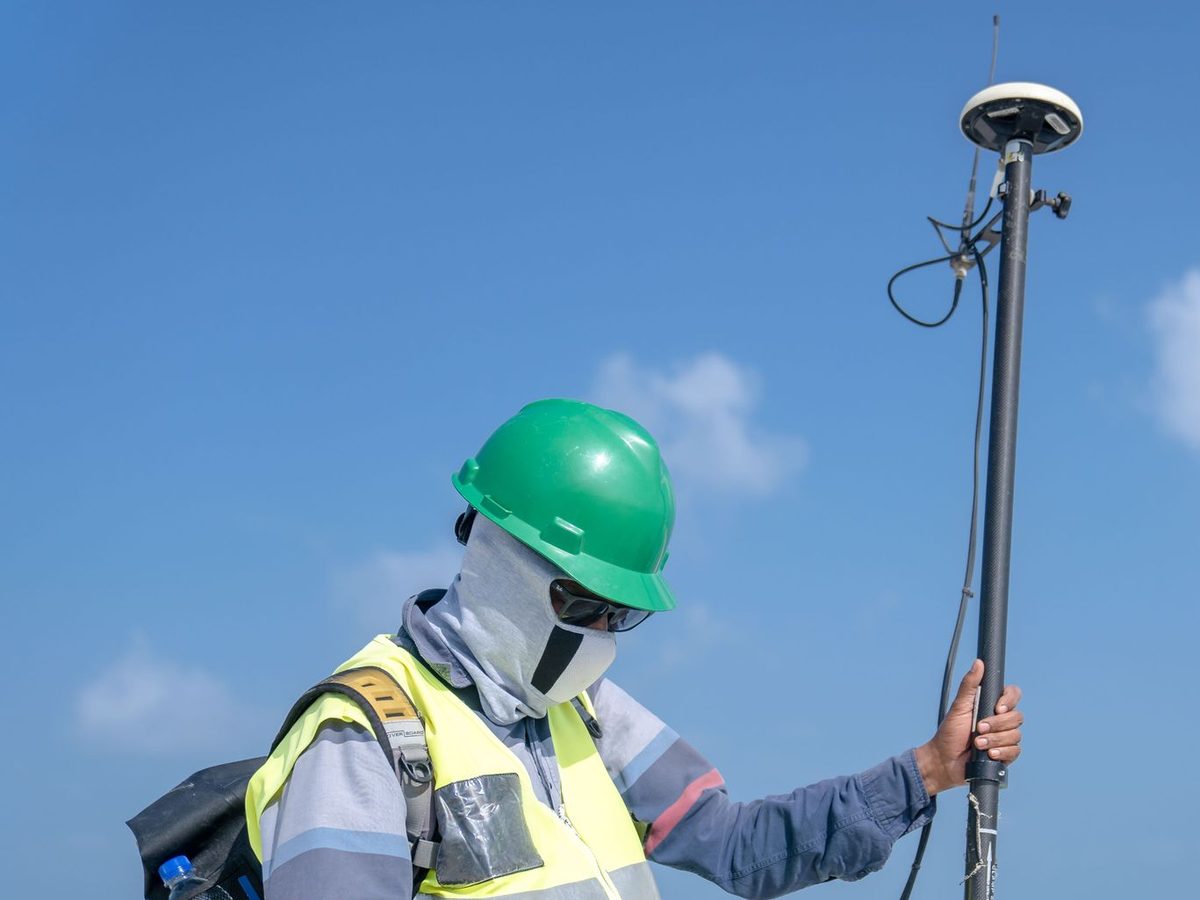 A worker in a green hard hat, goggles, face covering and yellow high-visibility vest holds a survey pole topped with a GNSS receiver antenna against a blue sky