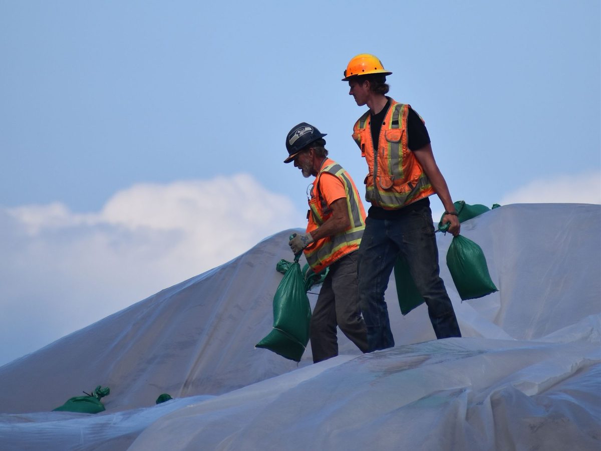 Two construction workers in hard hats and orange high-visibility vests carry green sandbags across white plastic sheeting under a blue sky