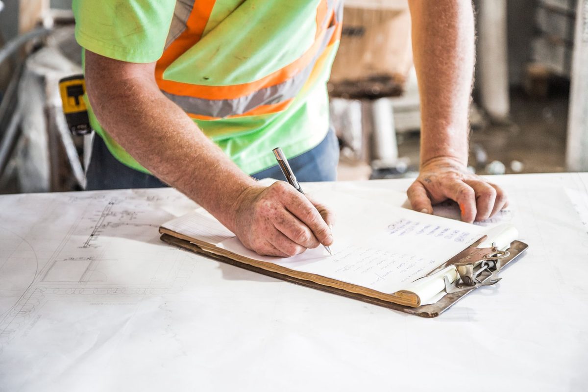 Close-up of a worker in a high-visibility vest writing on a clipboard that rests on a large sheet of plans