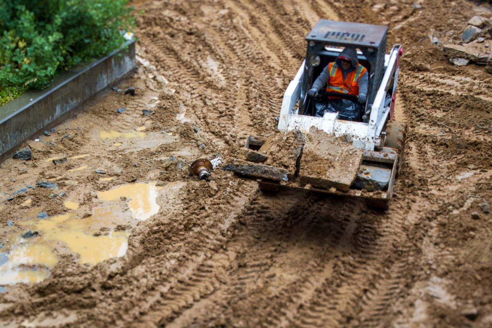 Skid-steer loader carrying broken concrete across a muddy, rutted construction site with puddles of standing water