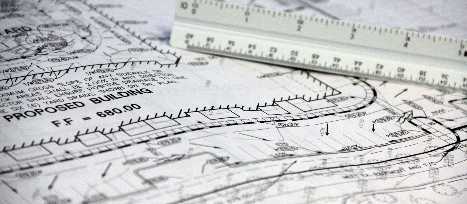 Close-up of a printed site grading plan showing spot elevations, drainage arrows and a proposed building pad, with a white triangular engineer's scale lying across it