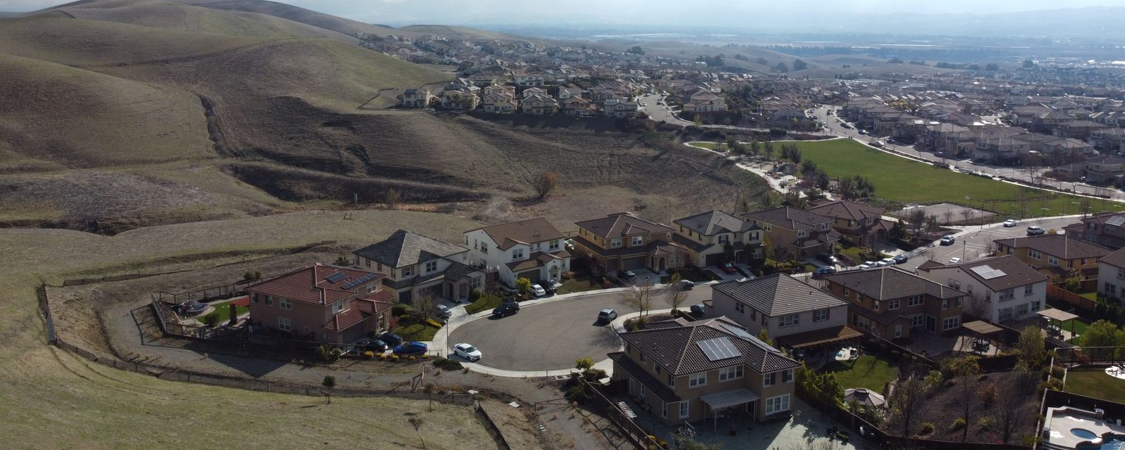 Aerial view of a hillside housing subdivision: two-story homes around a cul-de-sac, with dry grassy hills and more neighborhoods behind.