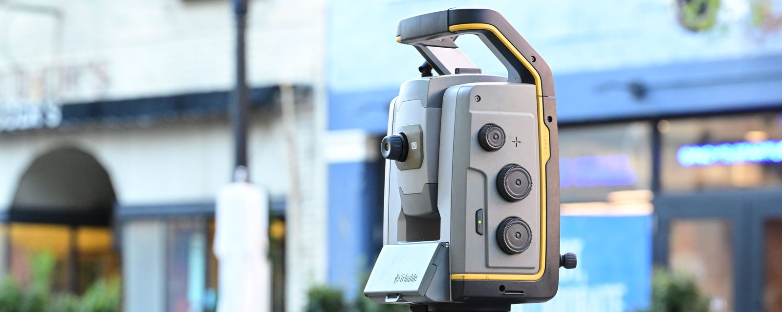 A robotic total station, seen close up, in front of blurred commercial storefronts and shrubs.