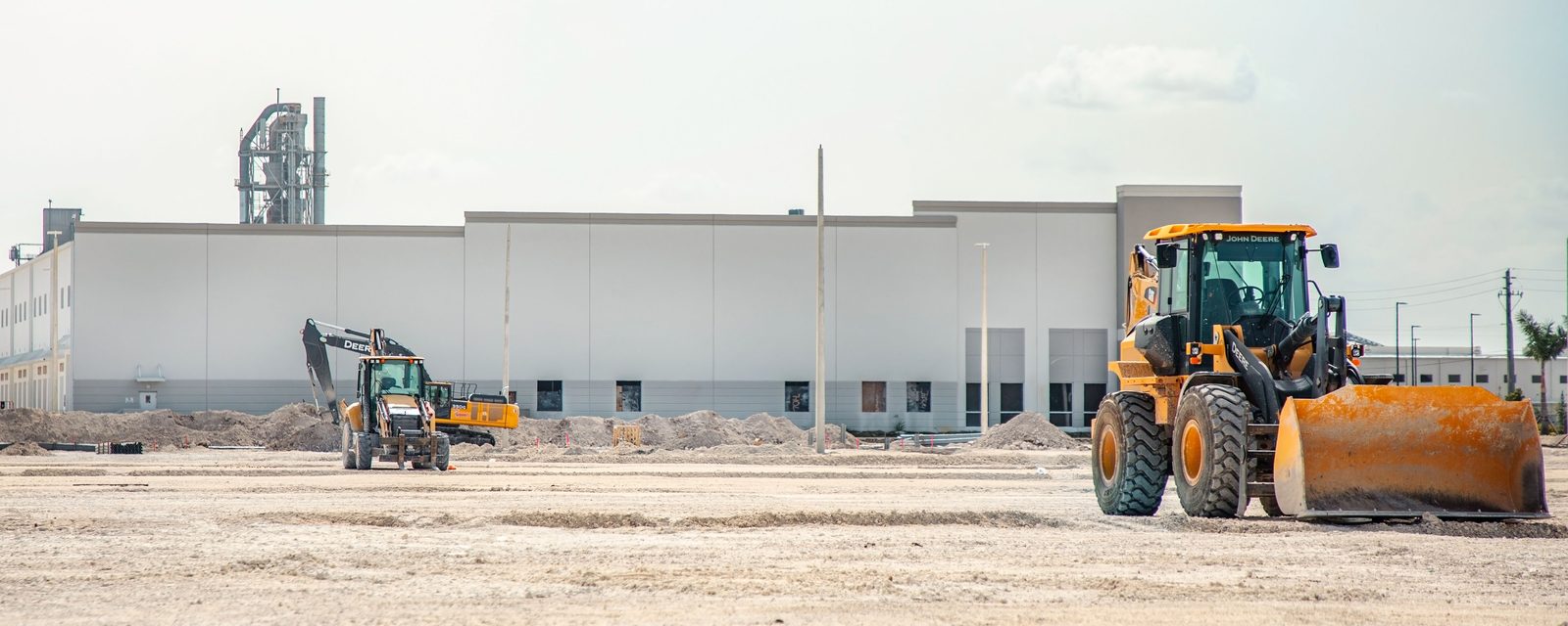 A yellow wheel loader, a backhoe loader, and an excavator on a large graded dirt pad with low soil piles, in front of a new white concrete-panel warehouse under a hazy sky.