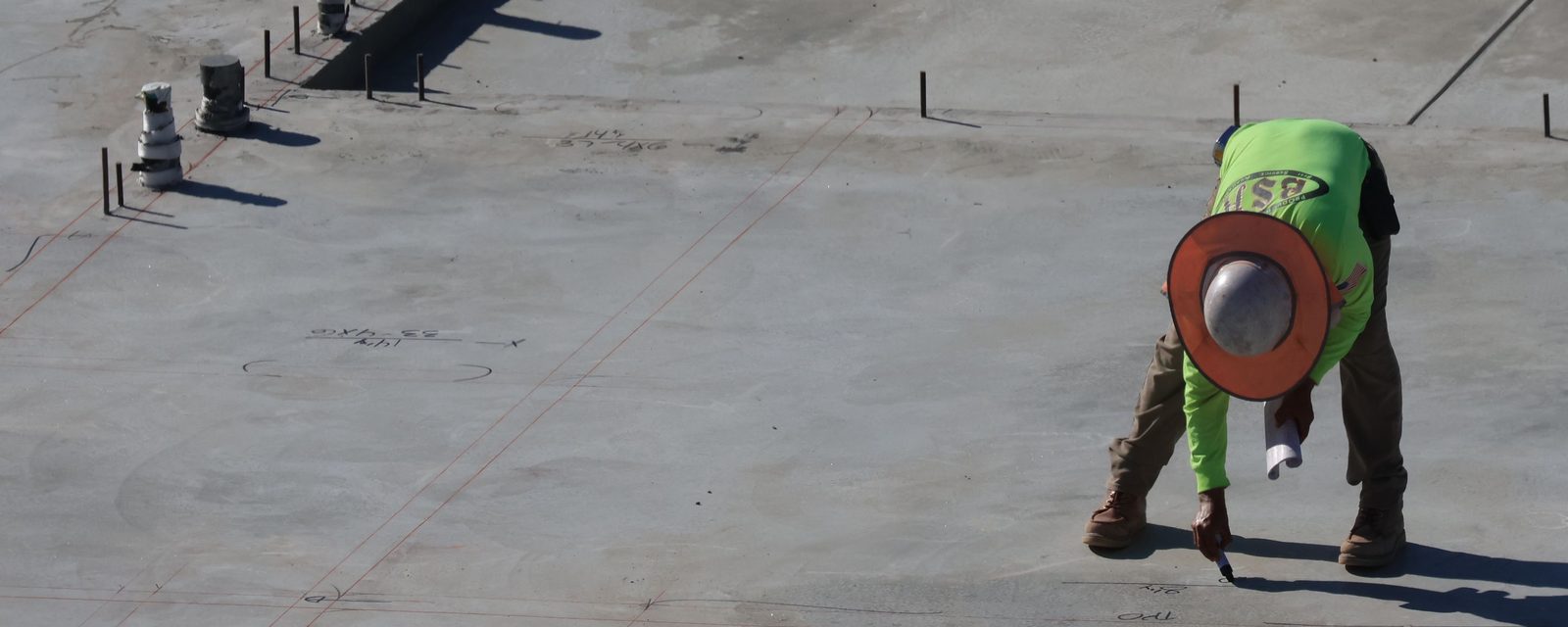 Seen from above, a worker in a hard hat and hi-vis shirt, holding rolled plans, writes a mark beside chalked layout lines on a new concrete slab.