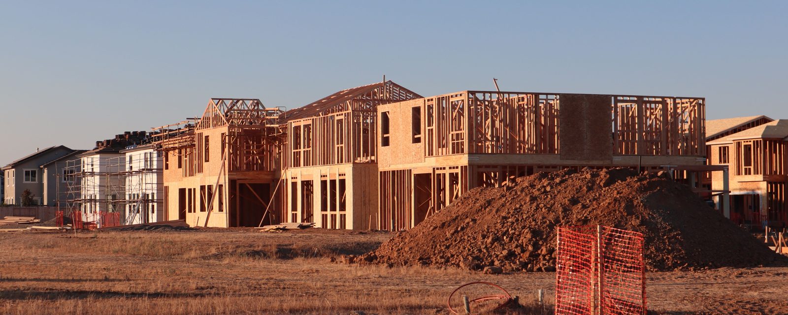 Wood-framed two-story homes under construction in a new housing development at sunset, with a large dirt pile and orange safety fencing in the foreground.