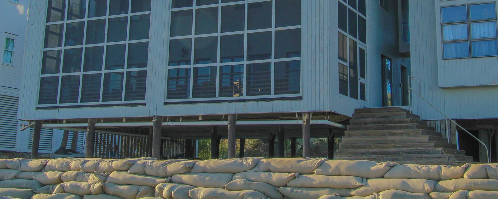 A beach house raised on wooden pilings above the sand, with wide wooden steps up to its main floor and a row of stacked sandbags along its front.