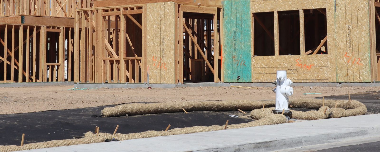 Straw fiber rolls staked along the edge of a new sidewalk and coiled around a white fire hydrant, in front of wood-framed homes under construction