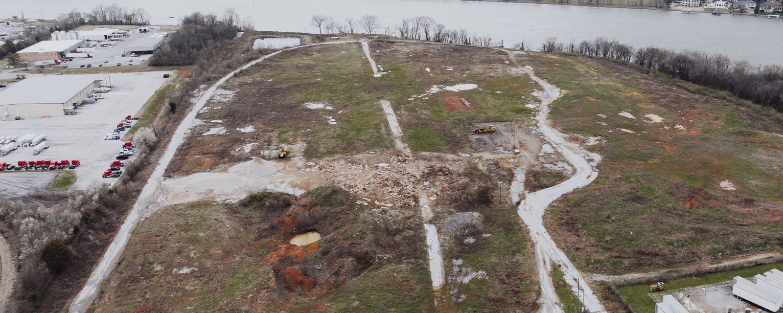 Aerial view of a large vacant parcel of patchy grass and bare dirt crossed by gravel roads, between warehouses and a wide tree-lined river.