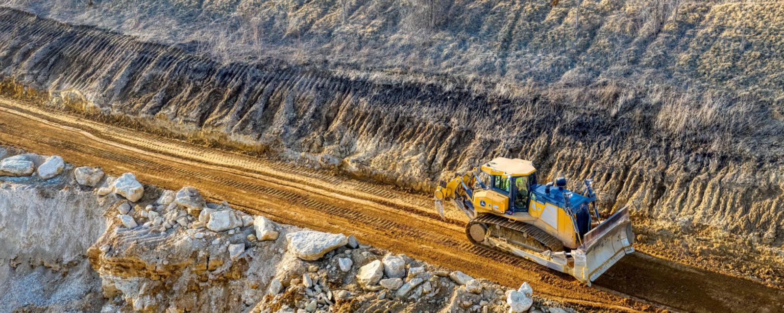 A yellow crawler bulldozer pushes dirt along a freshly cut bench at the foot of a graded earth slope, seen from above, with broken rock in the foreground.