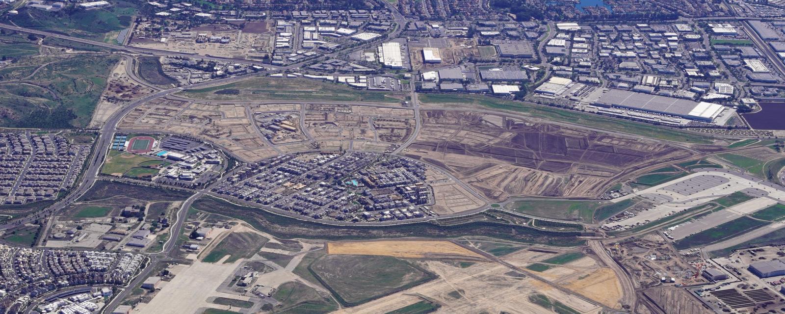 High-altitude aerial view of a large development area: freshly graded land and newly laid-out streets beside a new residential neighborhood, with industrial buildings beyond.