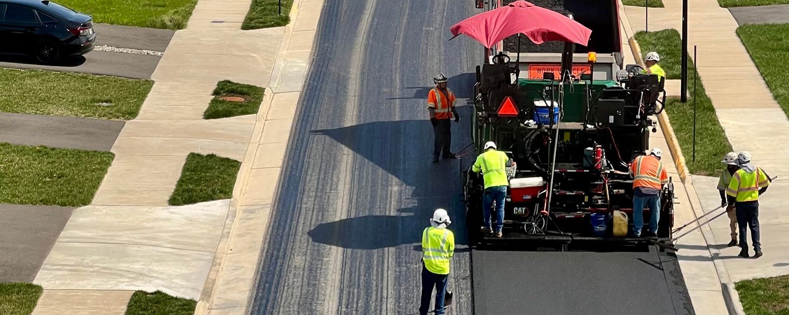 Overhead view of a residential street being paved between new curbs and sidewalks, with an asphalt paver, a dump truck and workers in hi-vis vests and hard hats.