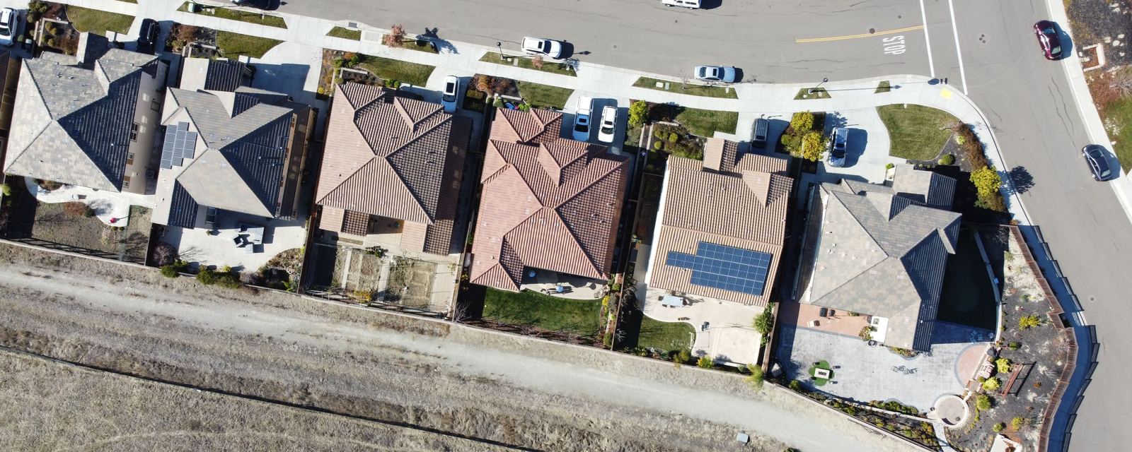 Overhead aerial view of a row of suburban houses whose fenced backyards back onto a dirt path and an open dry-grass slope, with a curving street and parked cars along the front.