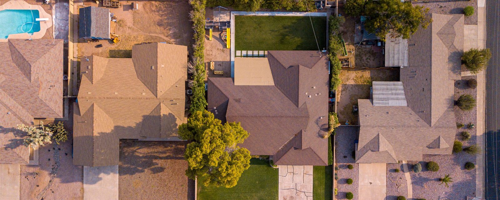 Aerial view looking straight down on three adjoining single-story houses, with hedges and walls along the shared lot lines between their yards, a front lawn, gravel landscaping and palm trees along the street.