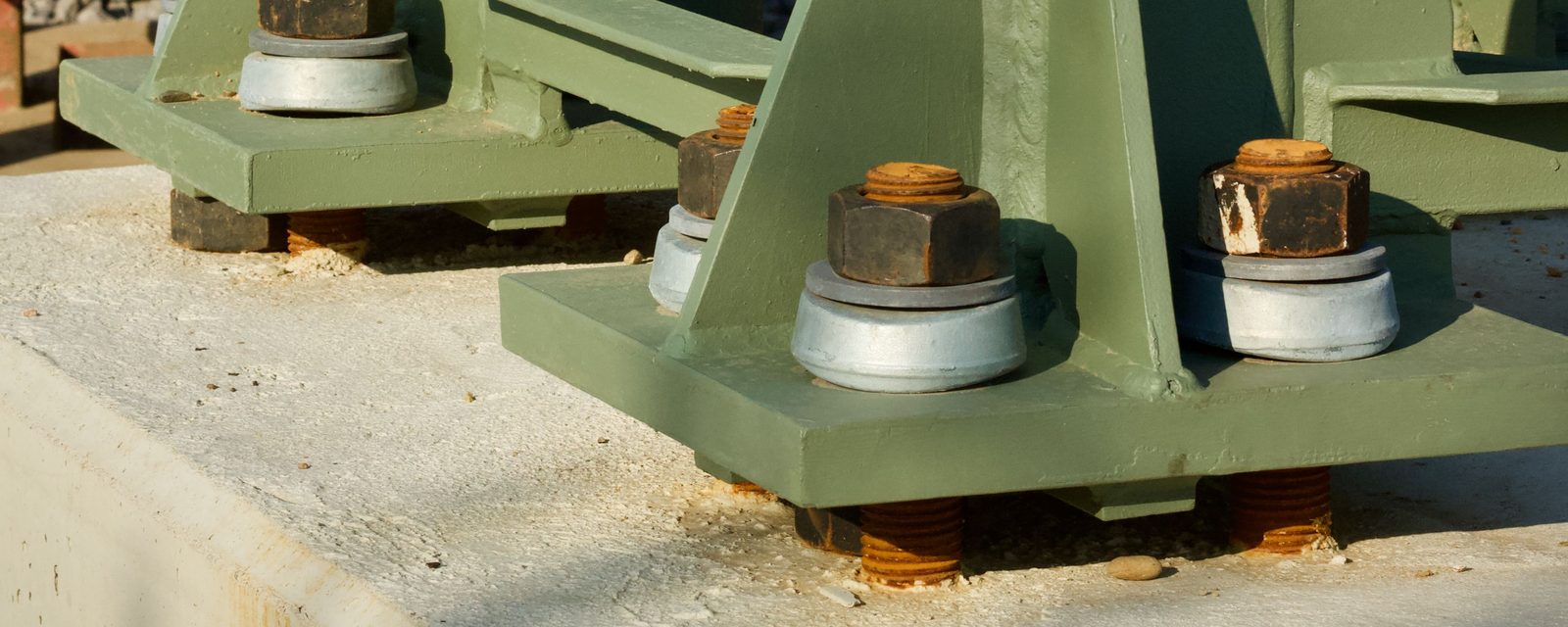 Close-up of two green steel column base plates bolted to a concrete foundation with hex nuts and washers on threaded anchor rods.