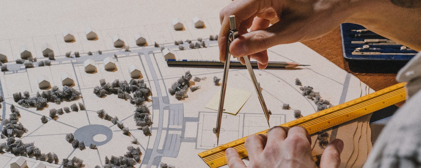 A person uses a drafting compass and a yellow scale ruler on a site-plan model of a residential neighborhood, with small model houses on lots, gray streets, foam trees and a traffic circle.