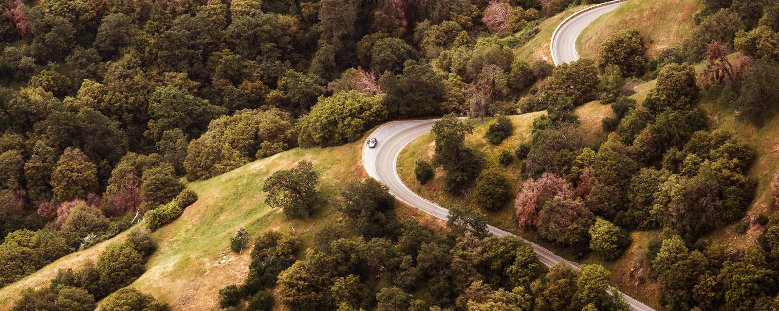 Aerial view of a narrow paved road winding in switchbacks through oak-covered foothills with grassy clearings.