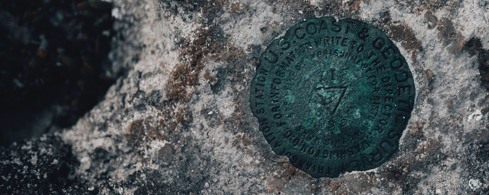 A weathered bronze U.S. Coast & Geodetic Survey triangulation station disk, green with age and stamped with a triangle, set flush in a gray rock outcrop.