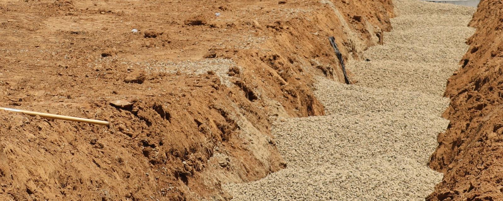 A trench bedded with light gray drain rock runs between walls of excavated reddish-brown soil on a graded dirt site.