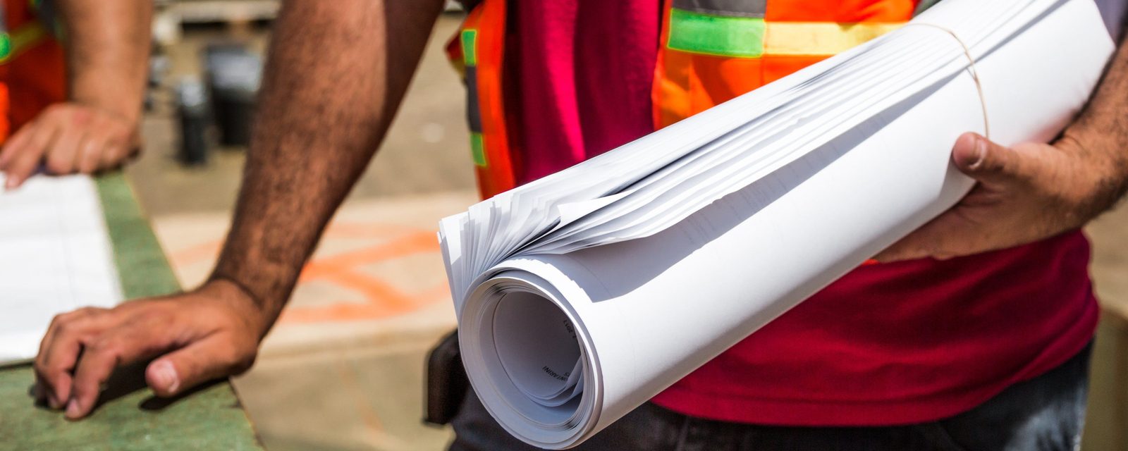 A worker in an orange high-visibility vest holds a thick roll of white plan sheets under one arm, beside a second person leaning on a table.